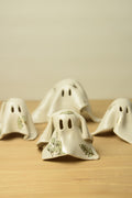 Four small ghost-shaped ceramic ornaments in off-white with green botanical leaf illustrations.