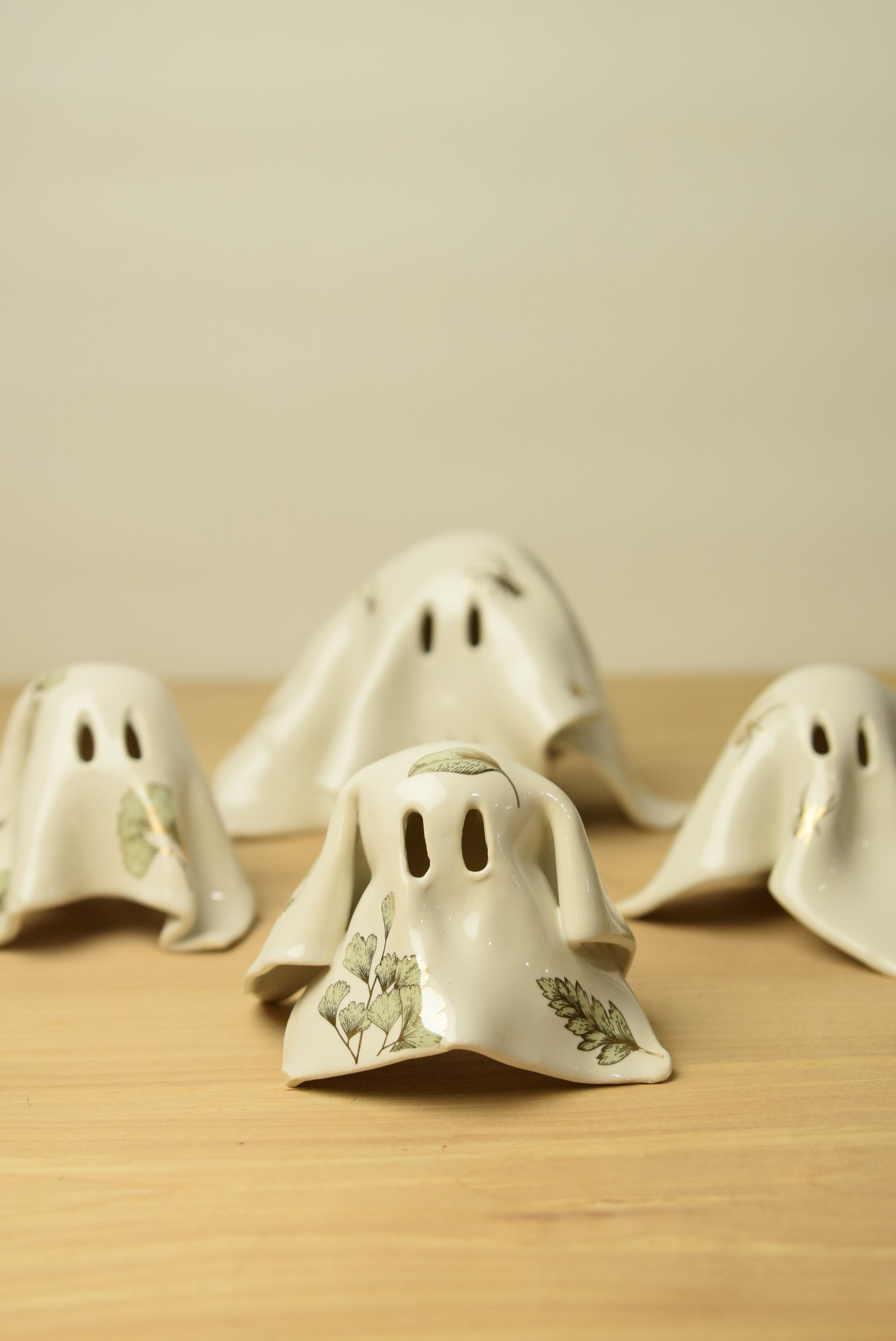 Four small ghost-shaped ceramic ornaments in off-white with green botanical leaf illustrations.